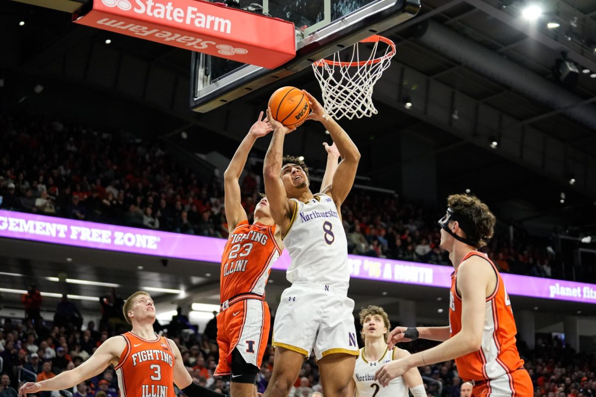 A basketball player jumps up toward the hoop, holding the ball in their raised arms, as an opposing player attempts to block them.