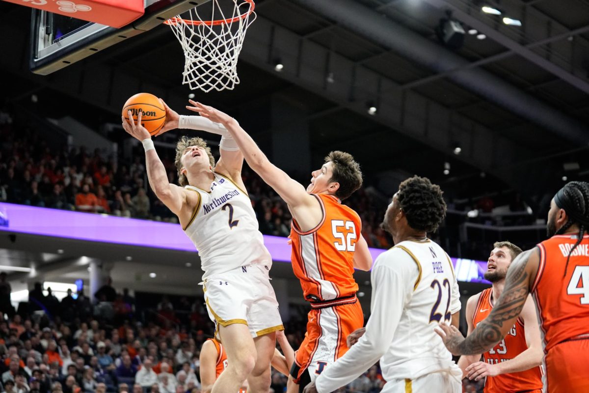 A basketball player jumps up toward the hoop, holding the ball in their raised arms, as an opposing player attempts to block them. Other players nearby watch.