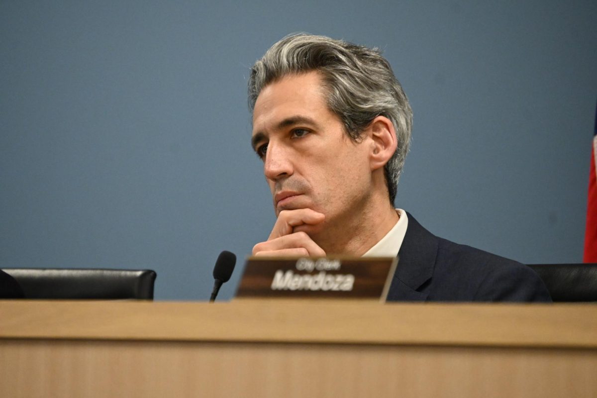 A person sitting behind a wooden table strokes their chin while staring at something off camera. A blurry nametag and mic are visible in front of them.