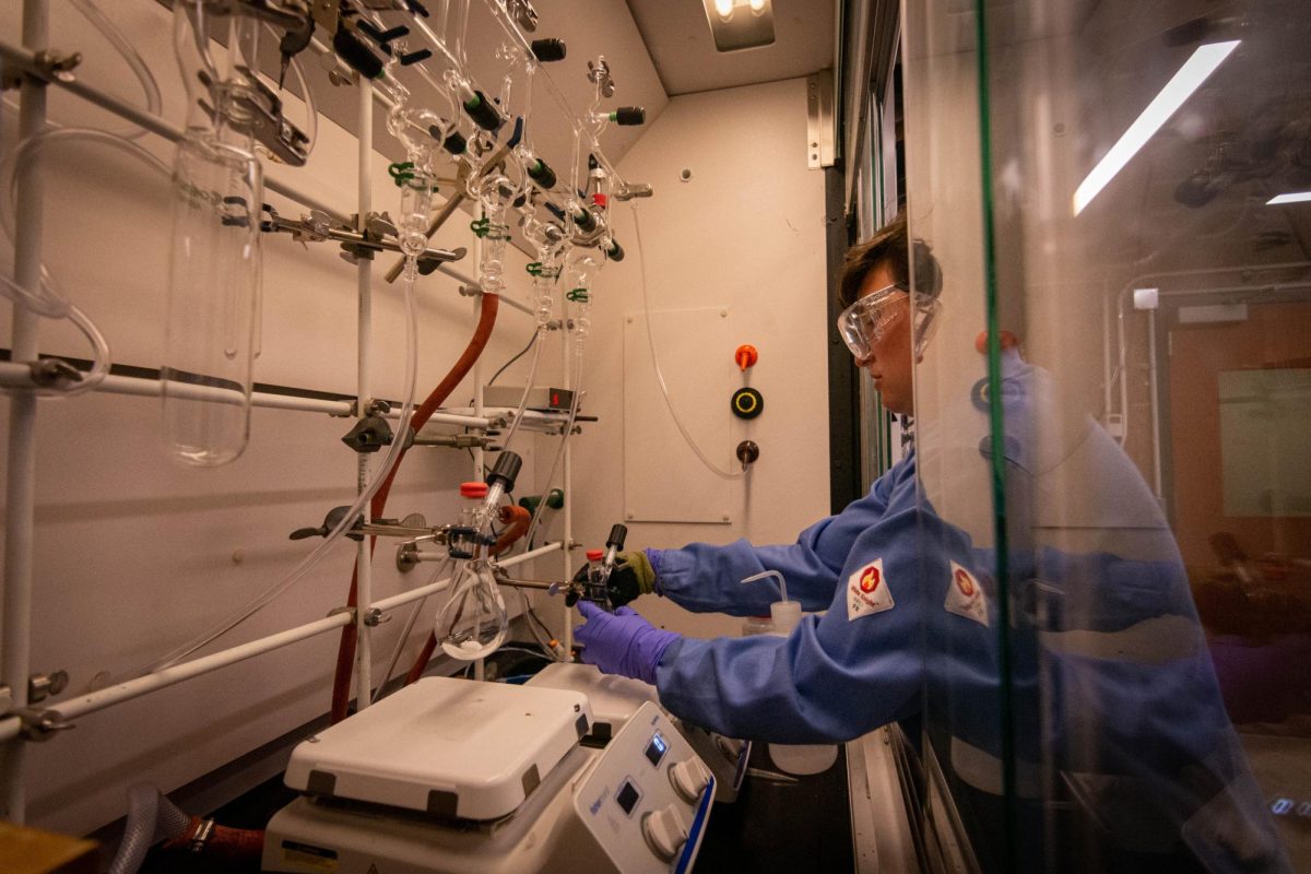 A student in a blue flame-proof lab coat with lab goggles handles solid matter in rounded flasks within a fume hood.