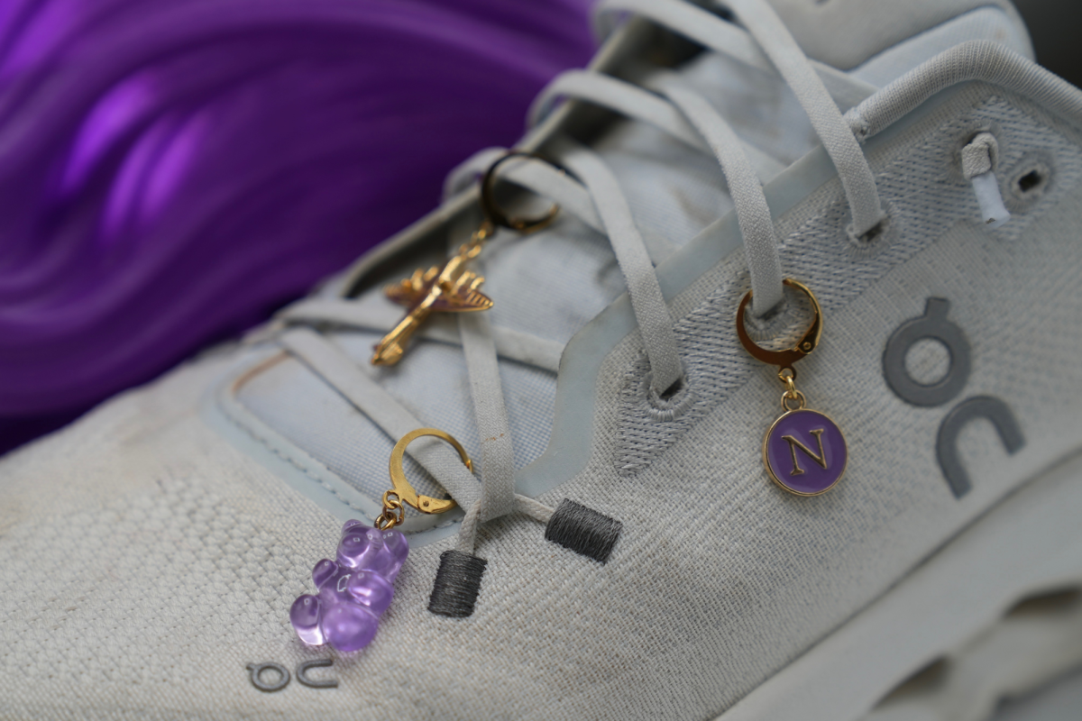 White sneakers are shown with charms of a purple teddy bear, a Northwestern “N” and a golden airplane are clipped onto the laces.