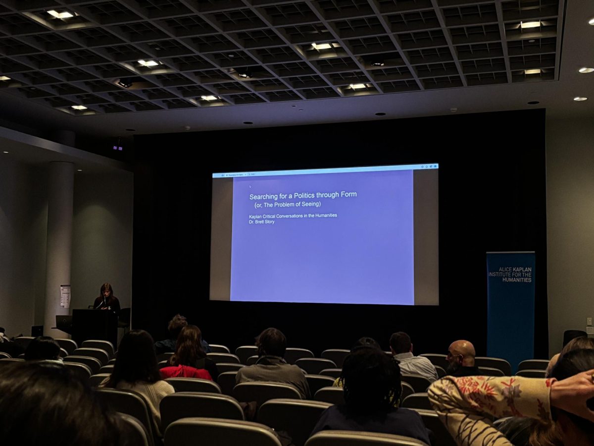 A woman presents to an audience at a podium next to a large slideshow projection.