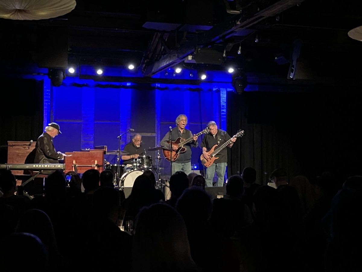 Four people play guitar, drums and a keyboard on stage.