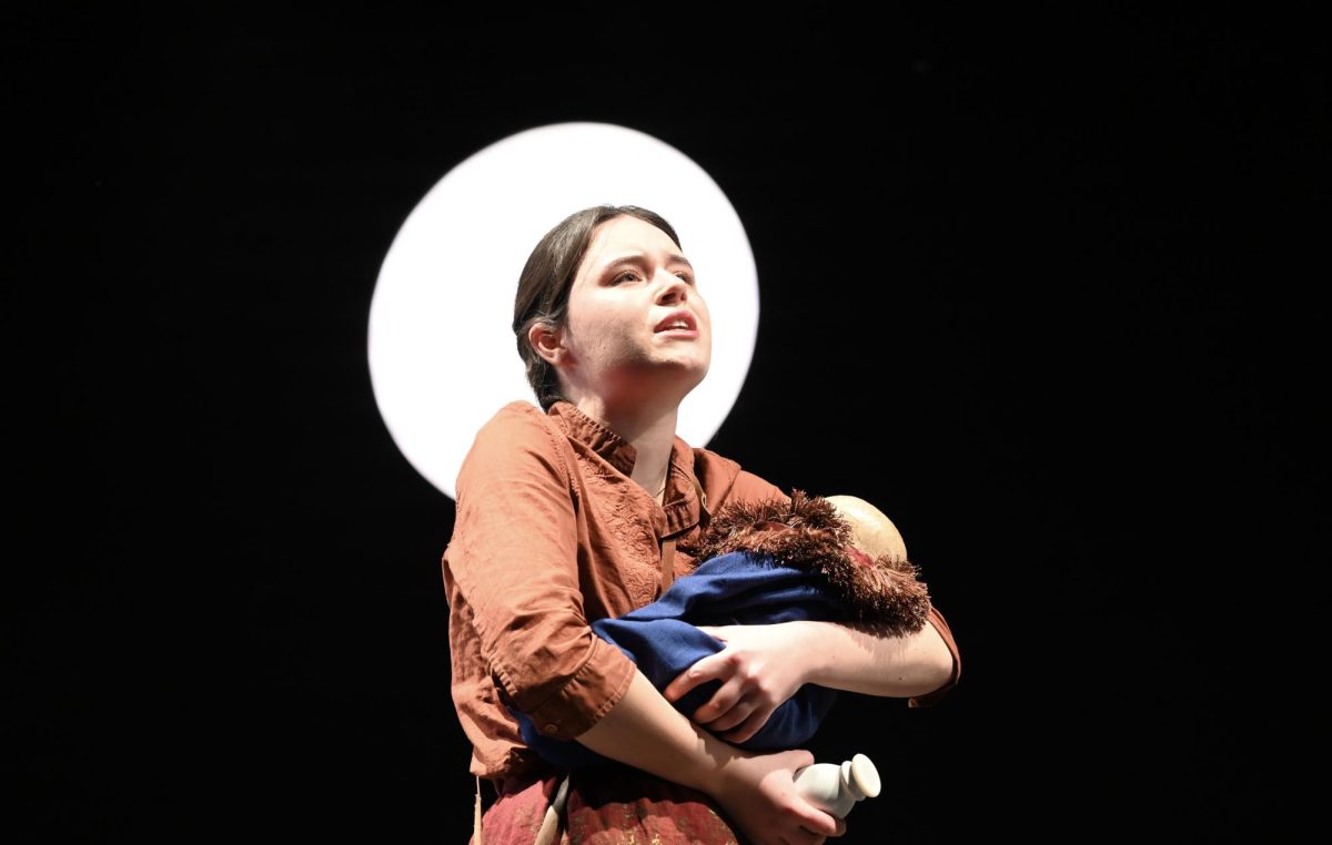 An actor holds a fake baby on stage in front of a white circle light.