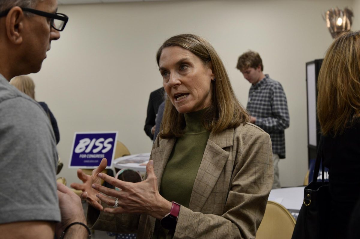 A person speaks with another person. Behind the person is a table with a “Biss for Congress” sign.
