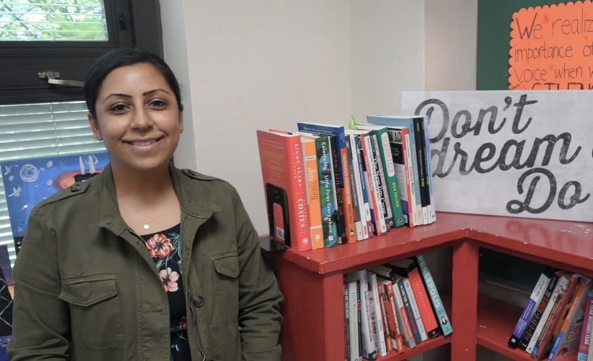 A person looks at the camera, smiling, while standing next to a bookshelf.