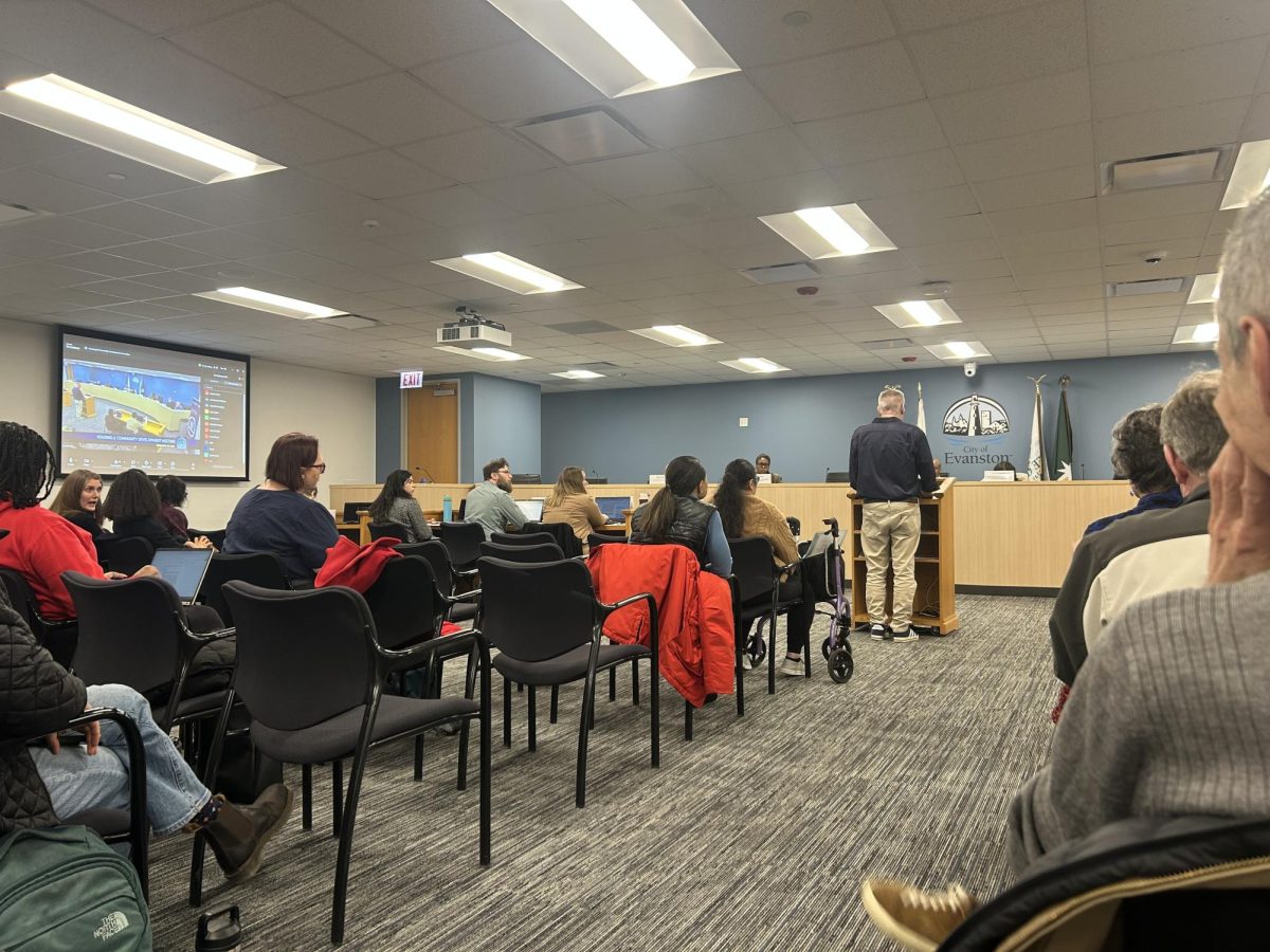 Evanston City Council Chambers full with people, person standing at podium at front of room in front of board members.