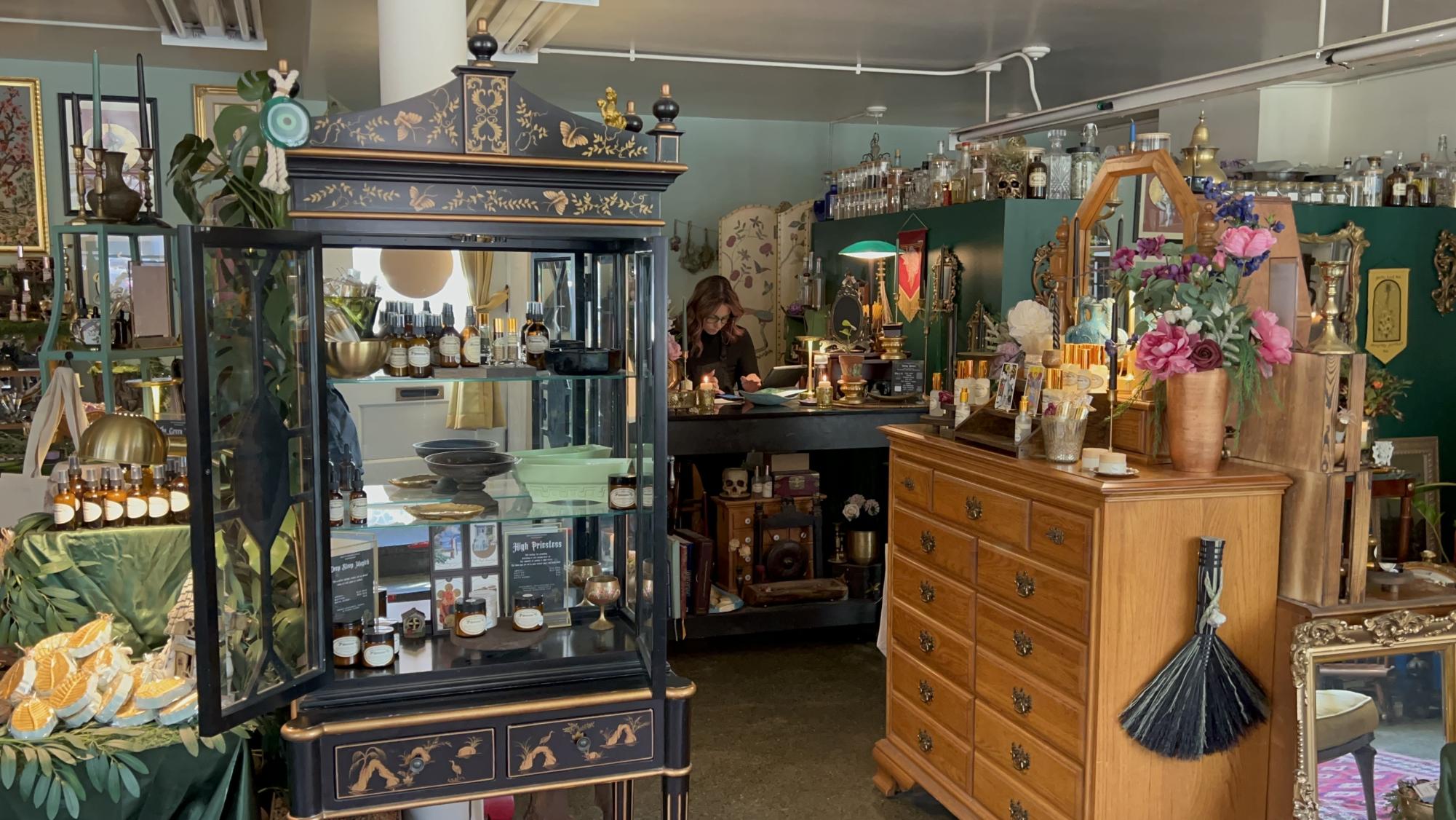 A black cabinet with floral engravings is filled with oil spray bottles. The background has various bottles, candles and plants clustered on different desks and on top of the wall.