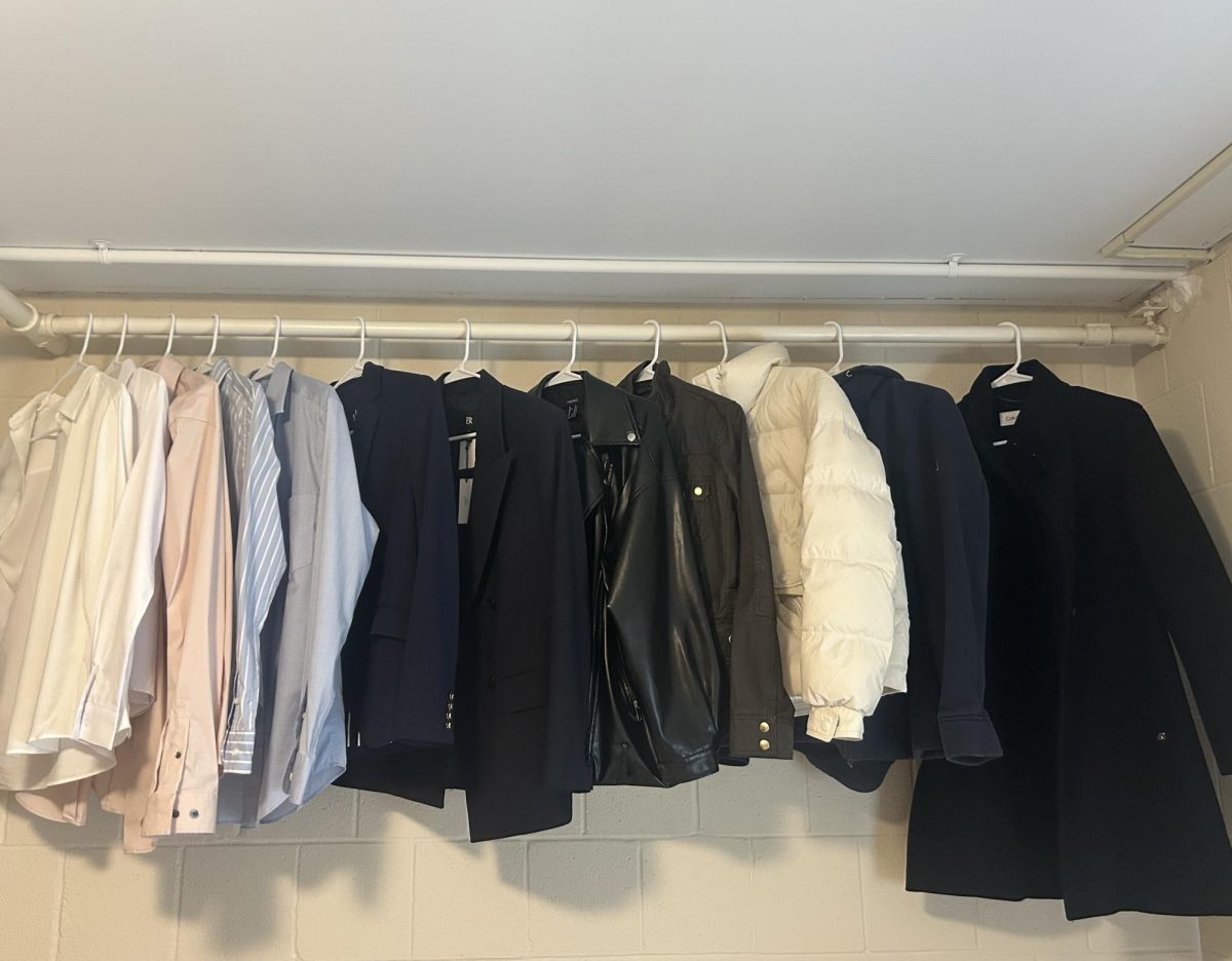 Neutral-toned shirts, navy and black blazers, leather jackets and a white puffer coat hang neatly on white hangers inside a dorm room closet.