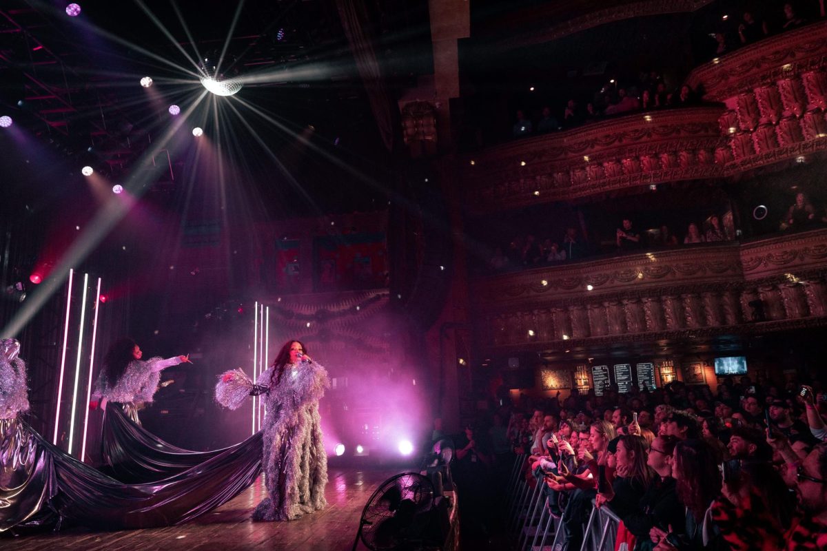 Three performers wearing extravagant silvery outfits dance onstage in front of a large crowd.
