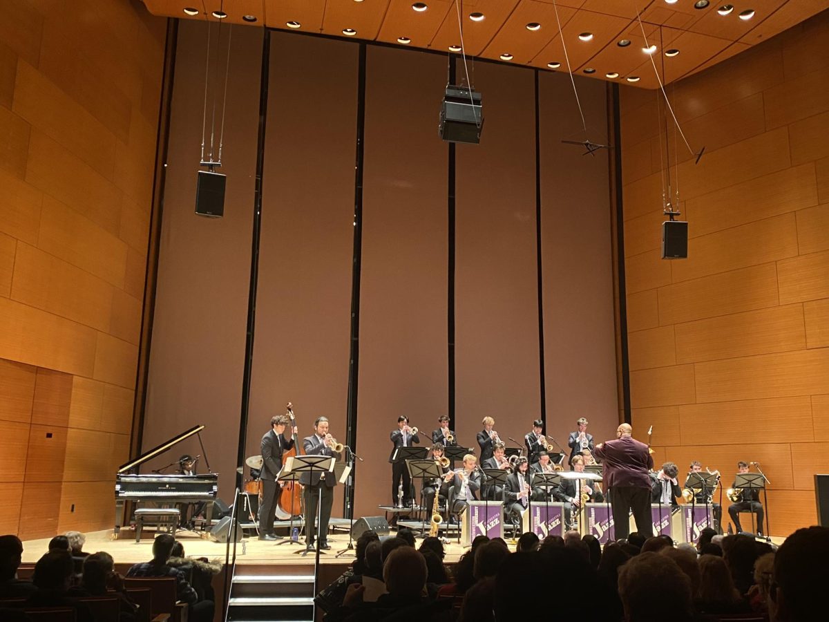 A person conducts people playing instruments on a stage. Other people sit in the audience.