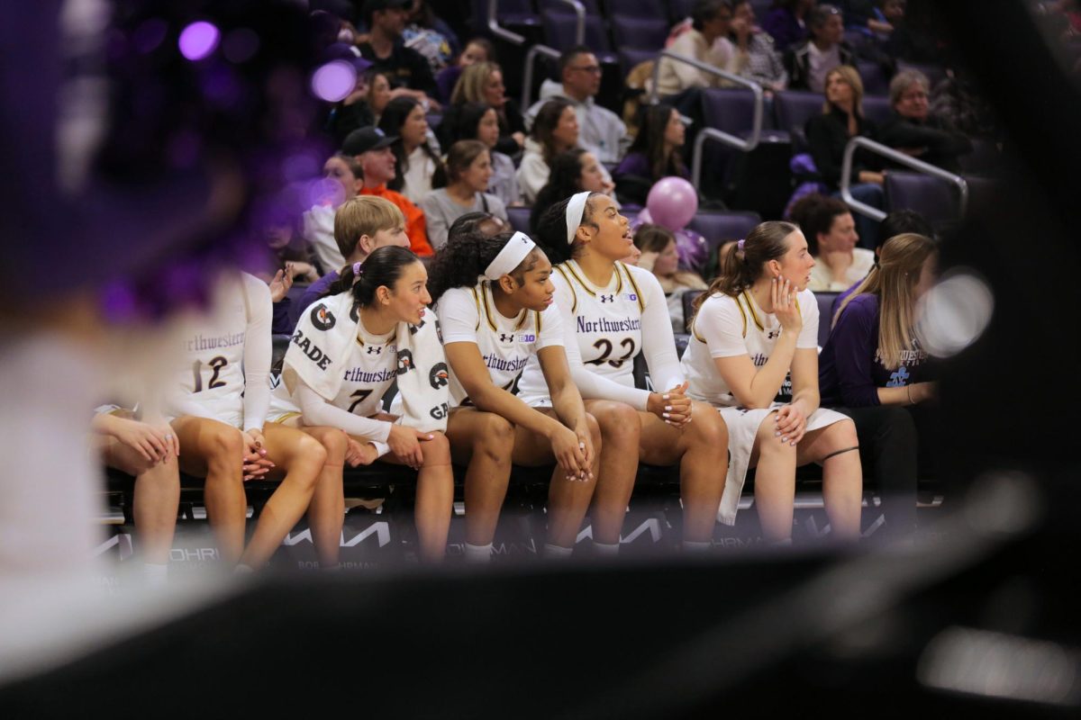 Several players in white jerseys sit on the bench and look at the court.