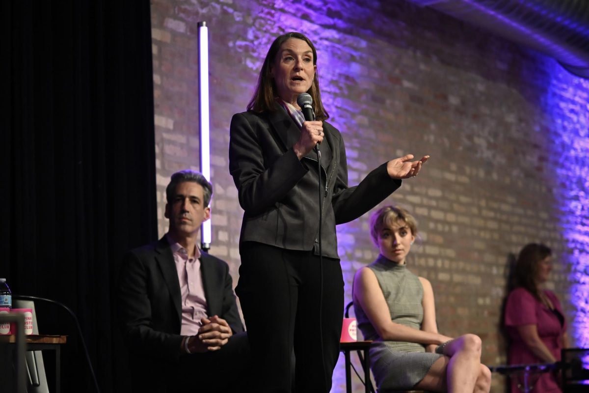 A person stands and speaks with a microphone at a campaign forum. Two people sit behind her.