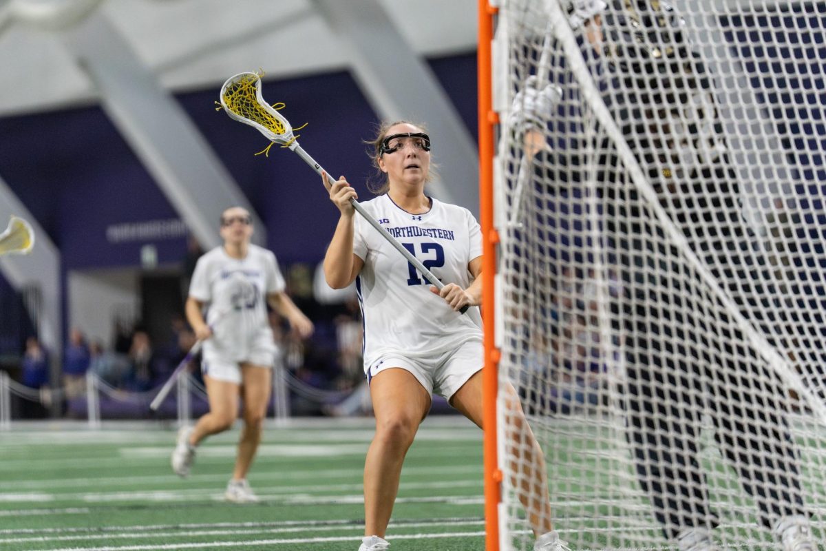 A lacrosse player stands in front of the opposing team&rsquo;s goal, holding their stick in the air.