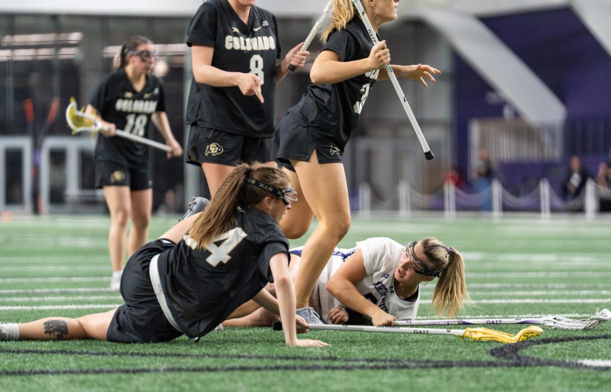 A lacrosse player lays on the ground after a fall, surrounded by opposing players, one of whom has also fallen.