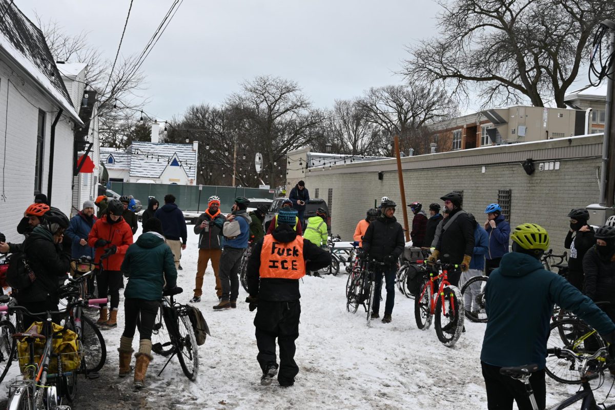 People stand with bicycles in the snow. One person has “F—k ICE” written on their back.