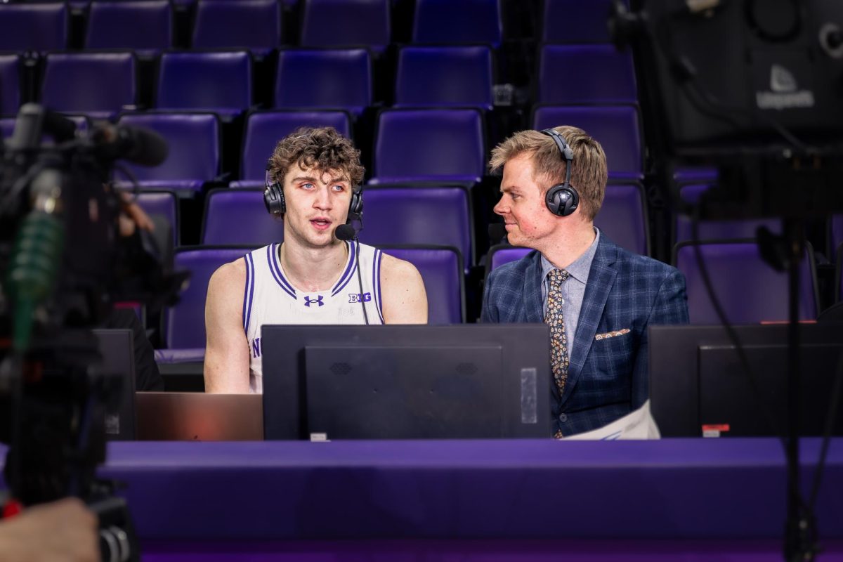 One basketball player and one person in a suit sit talking to each other with headphones on.