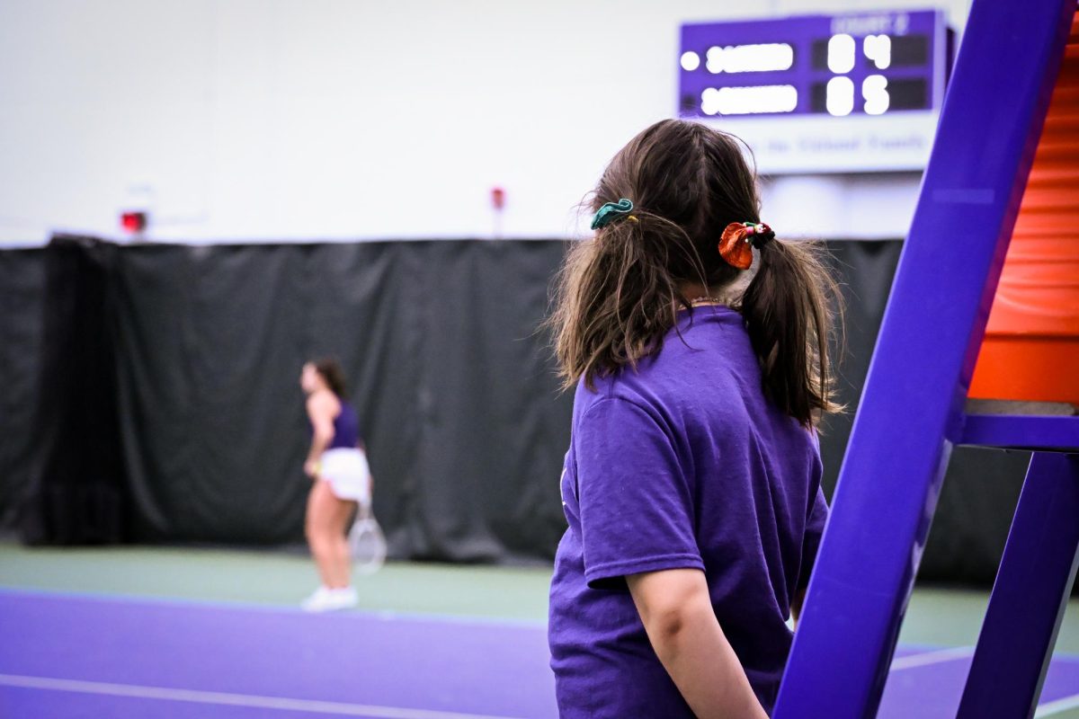 A child with pigtails watches a Northwestern tennis player serve. 
