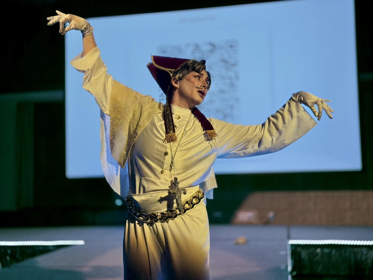 A drag king dressed with a pope costume, male makeup and short brunette wig contorts their arms.