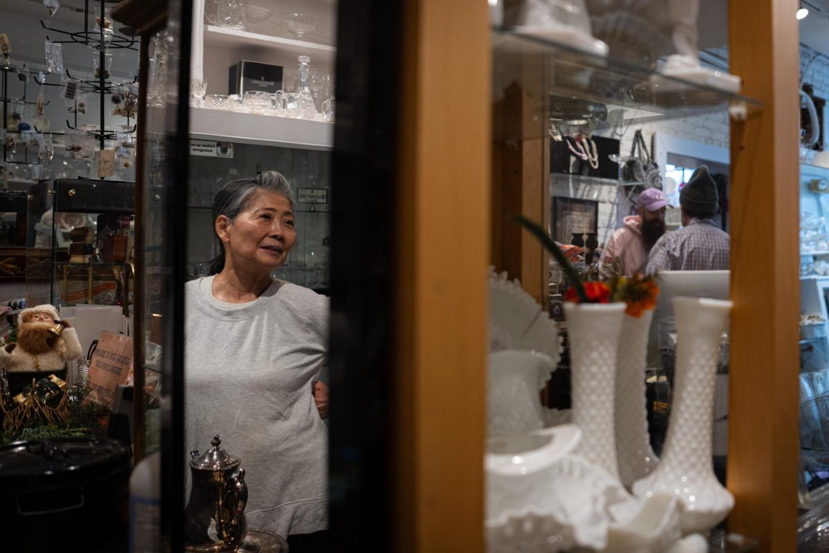 A woman is reflected in a mirror. Behind a shelf, two people talk to each other.