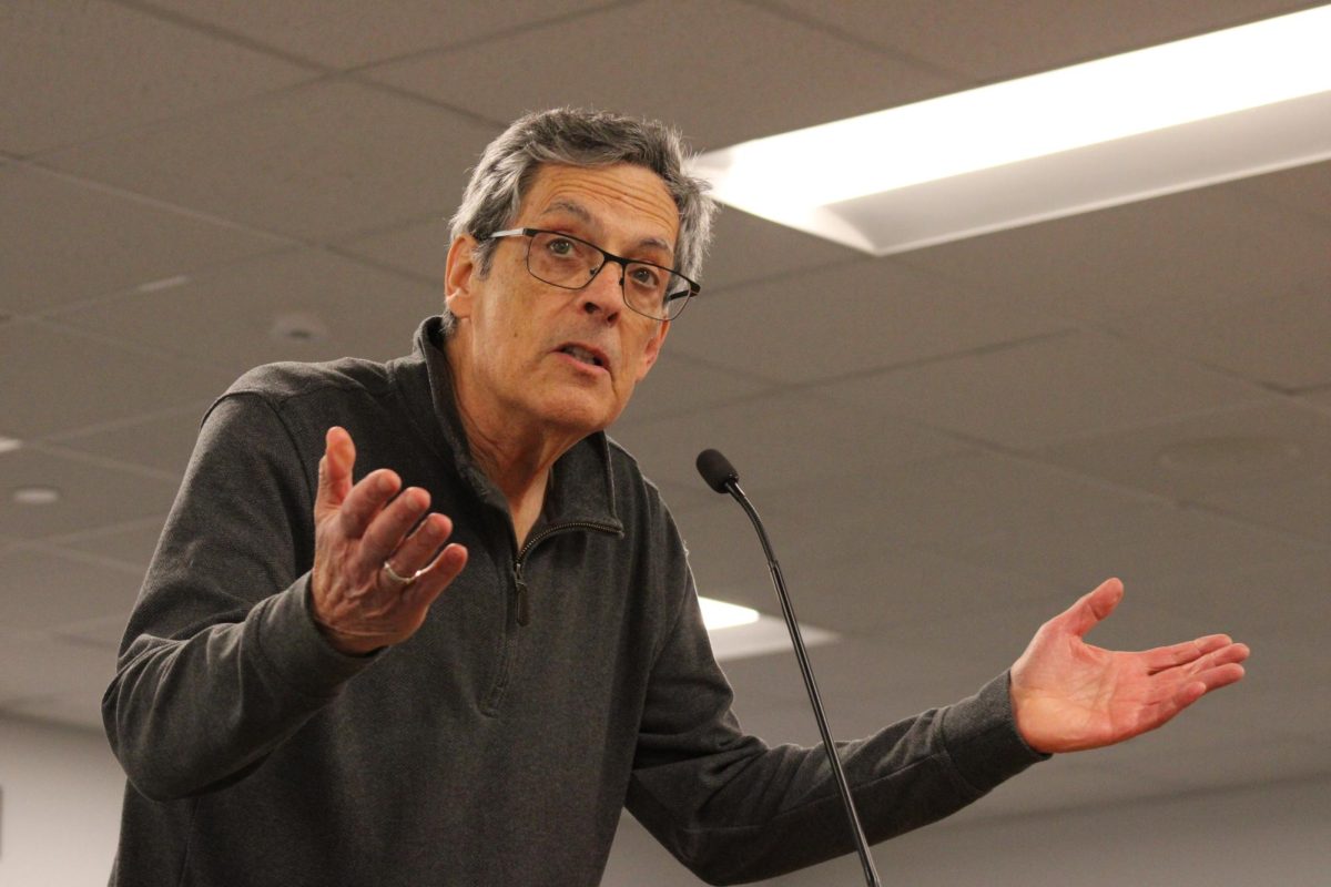A speaker in a grey sweater looks above the camera while speaking and making a shrugging gesture.