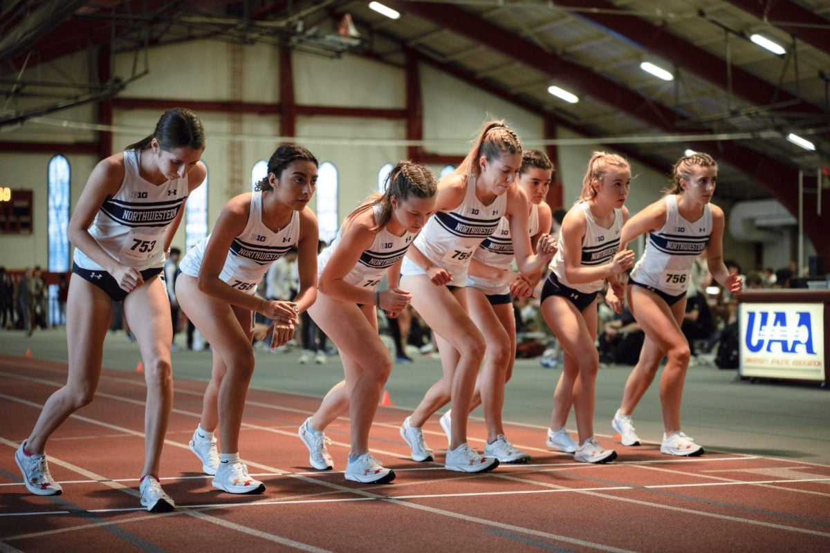 Seven runners in white uniforms line up at the starting line of an indoor track.