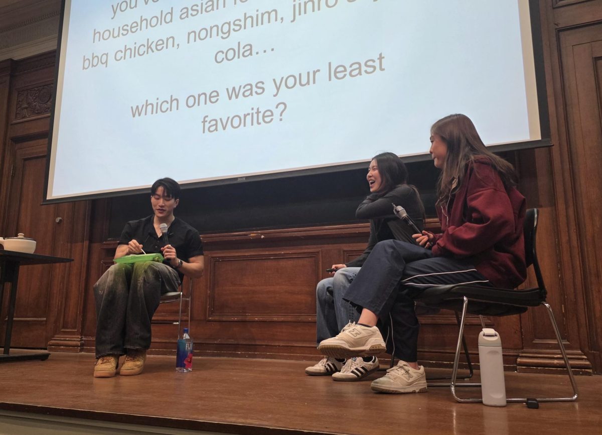 A man sits on a stage while eating. Two girls are smiling while sitting on the stage near the man.