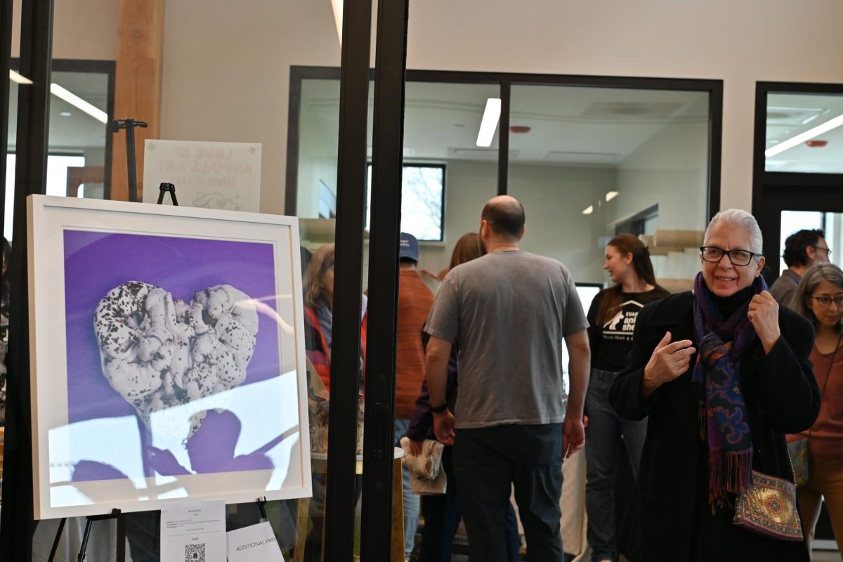 An art piece with white dogs creating a heart on a purple background. A person can be seen smiling while viewing the heart artwork. A crowd looking at other art is behind the dog photo.
