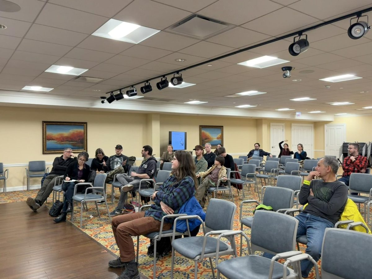 A small crowd sits in rows of chairs, with one person speaking, another raising their hand, one diligently taking notes and others listening intently.
