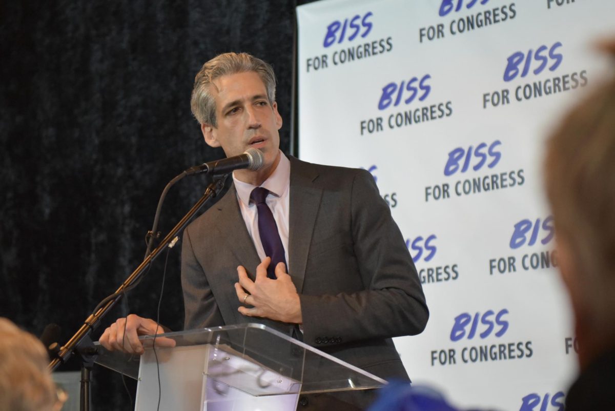 A person wearing a gray suit with a purple tie speaks into a microphone.