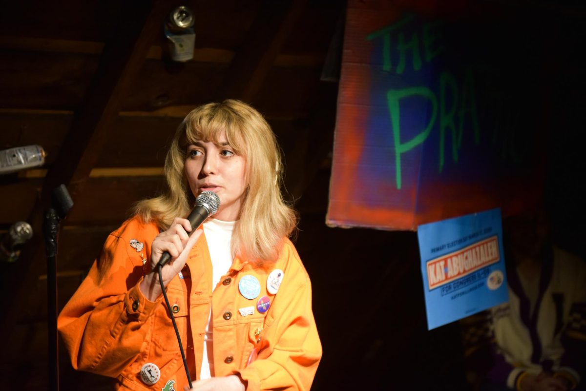 A person wearing an orange denim jacket with lots of pins speaks into a microphone.