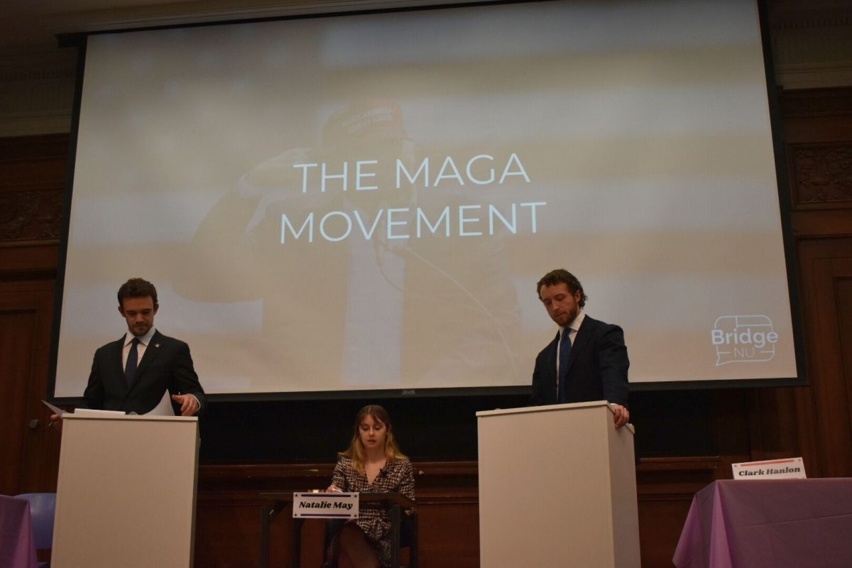 Two debaters stand at podiums as a moderator sits in a chair in between them.