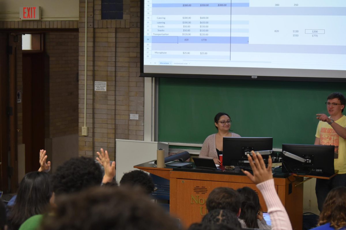 Students raise their hands, and two people stand at the front.