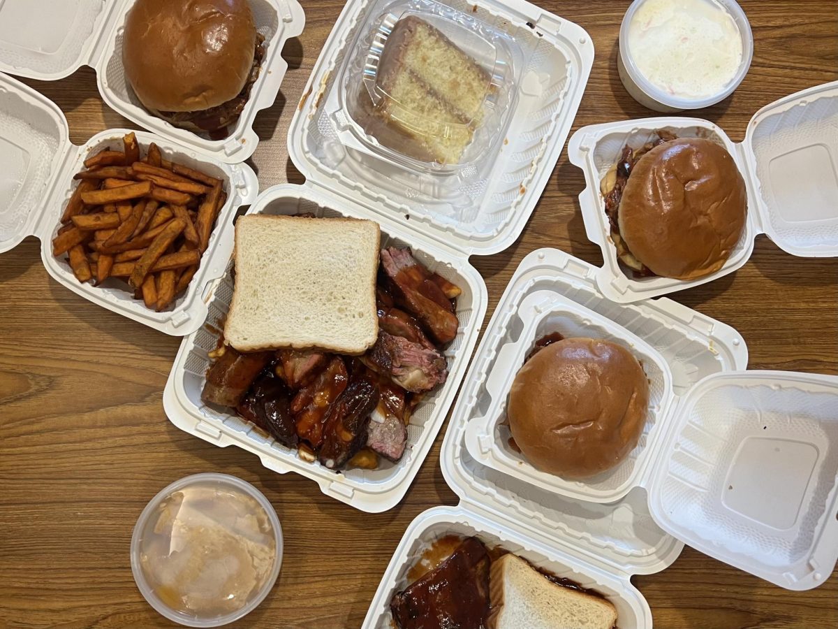 Three pulled pork sandwiches, a box of multiple kinds of meat, peach cobbler, a slice of cake and coleslaw on a table.