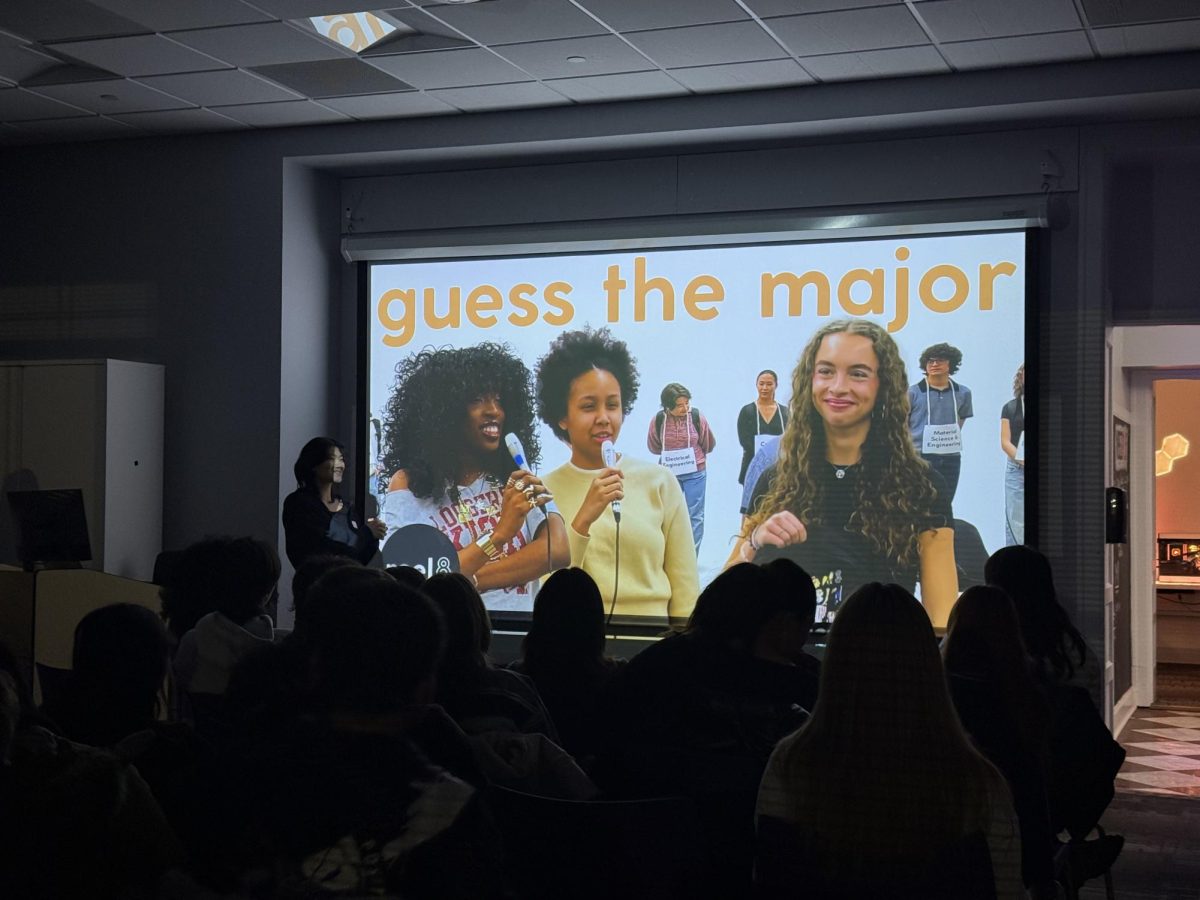 A person presents in front of an audience with a screen playing a video with the words “Guess the major.”