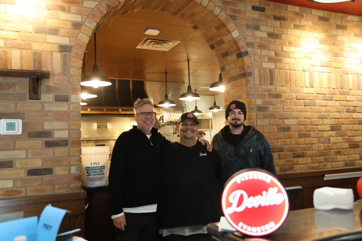Three people stand behind a lit-up Pizzeria DeVille sign on the counter.