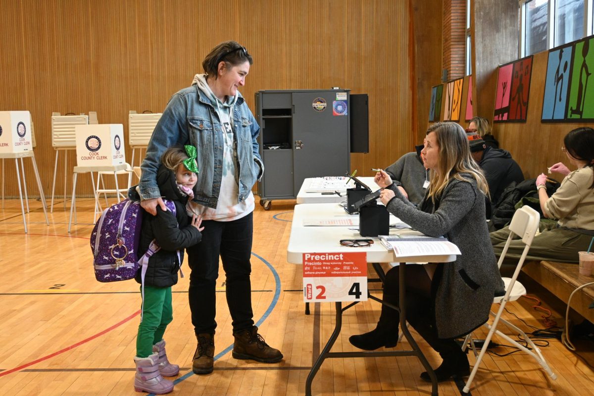 A parent with a child wearing a green bow stands in front of a table, speaking to a poll worker.