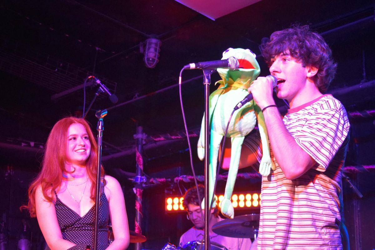 A performer holding a hand puppet sings into a microphone while another person to the side watches.