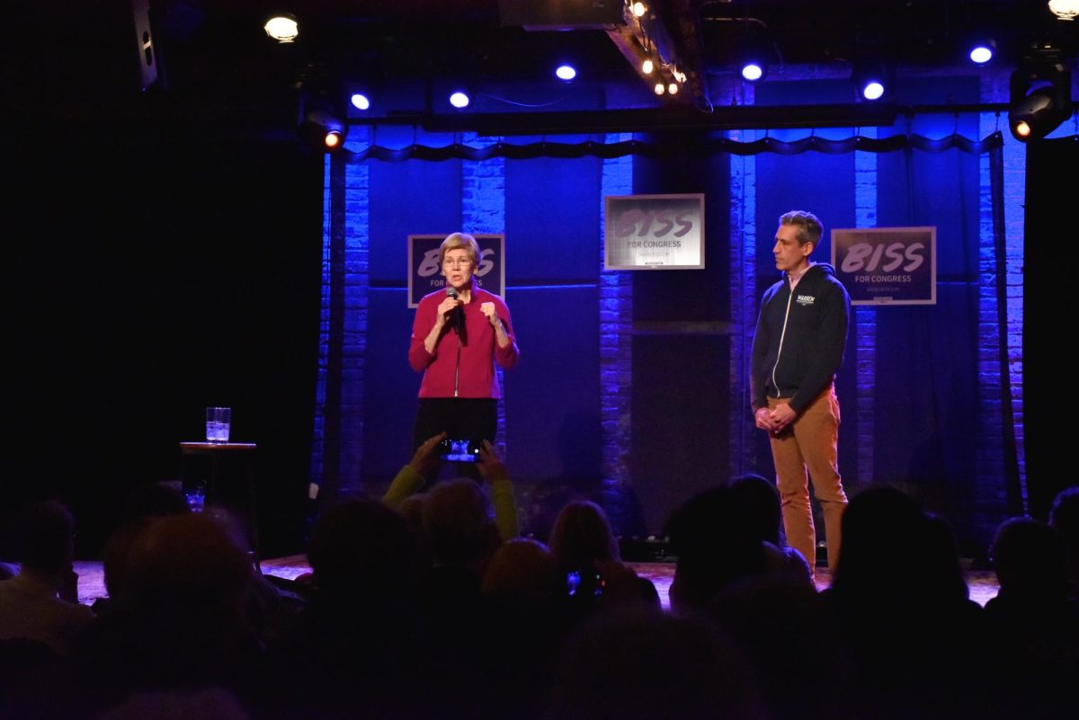 A person speaks into a microphone on a stage, with another person standing to their right.