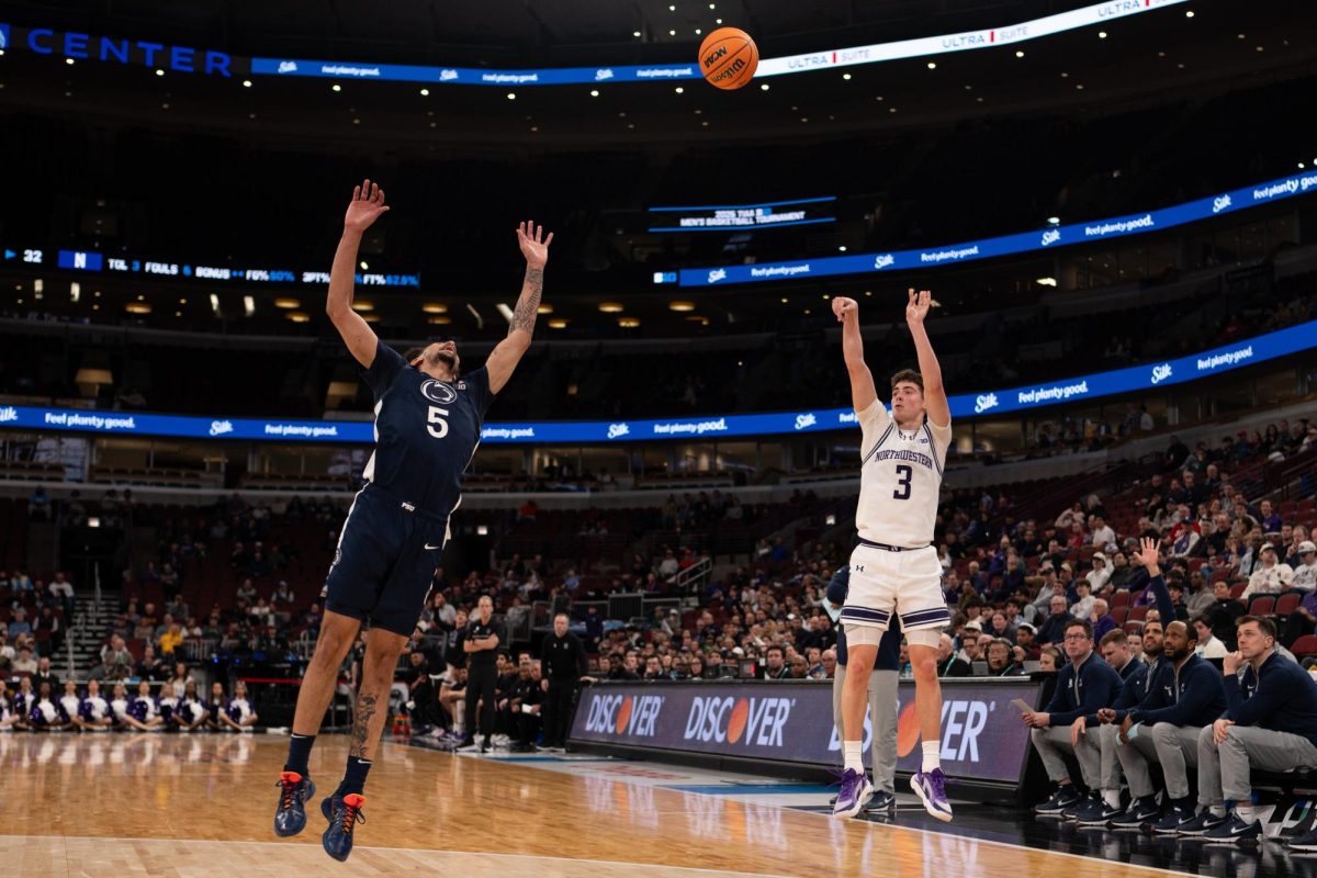 A player in a white jersey shoots a 3-pointer while a player in a blue jersey defends. 