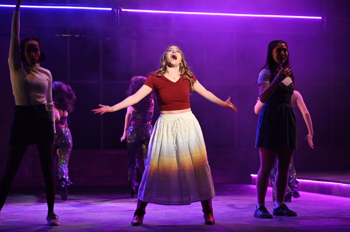 An actor wearing a red shirt and white skirt spreads their arms while singing, surrounded by other actors.
