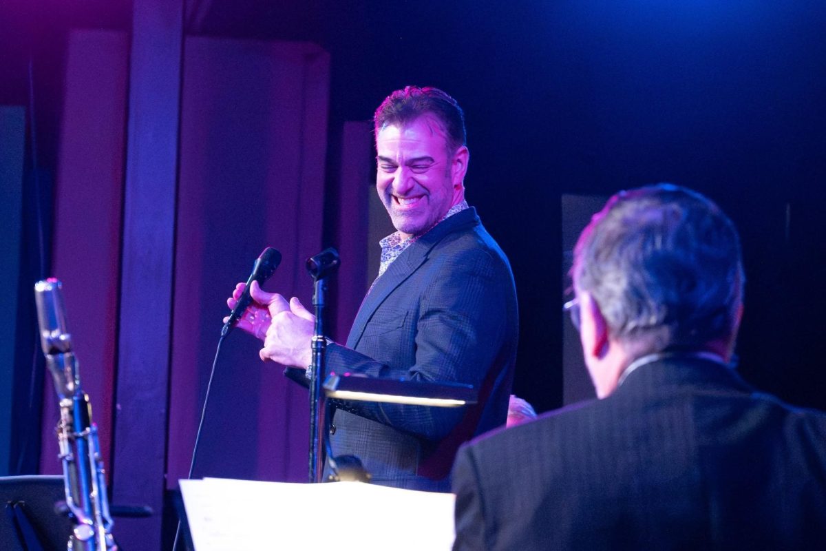 A person in a blue suit smiles down at somebody while holding a microphone. They&rsquo;re on a stage, bathed in blue and purple light.