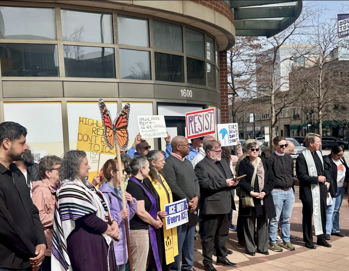 A crowd of people stand in front of a building labeled “1600.” Some hold cardboard signs.