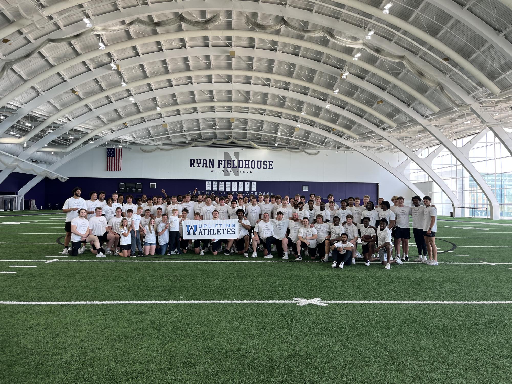 A large group of people in white shirts stands together on a football field, smiling for a photo and holding a white banner that reads “Uplifting Athletes.”