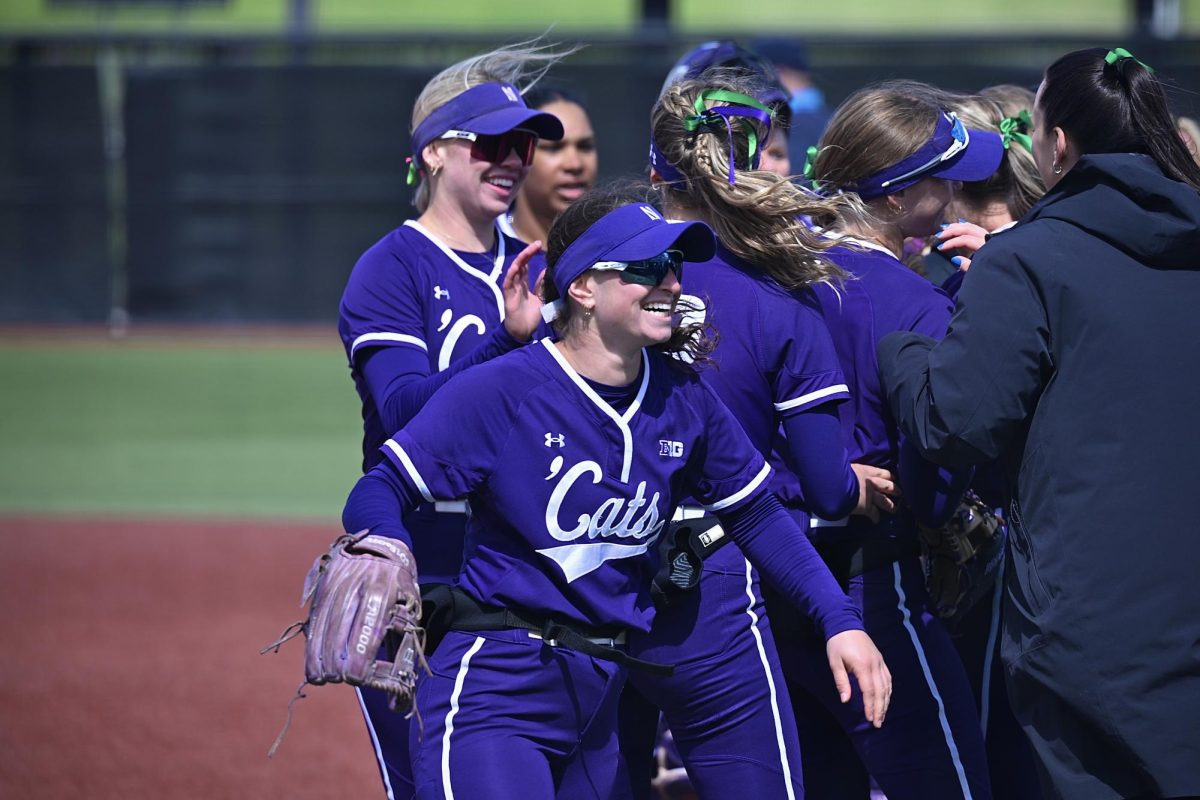 Northwestern softball players wearing purple jerseys smile and celebrate on the field after a win.