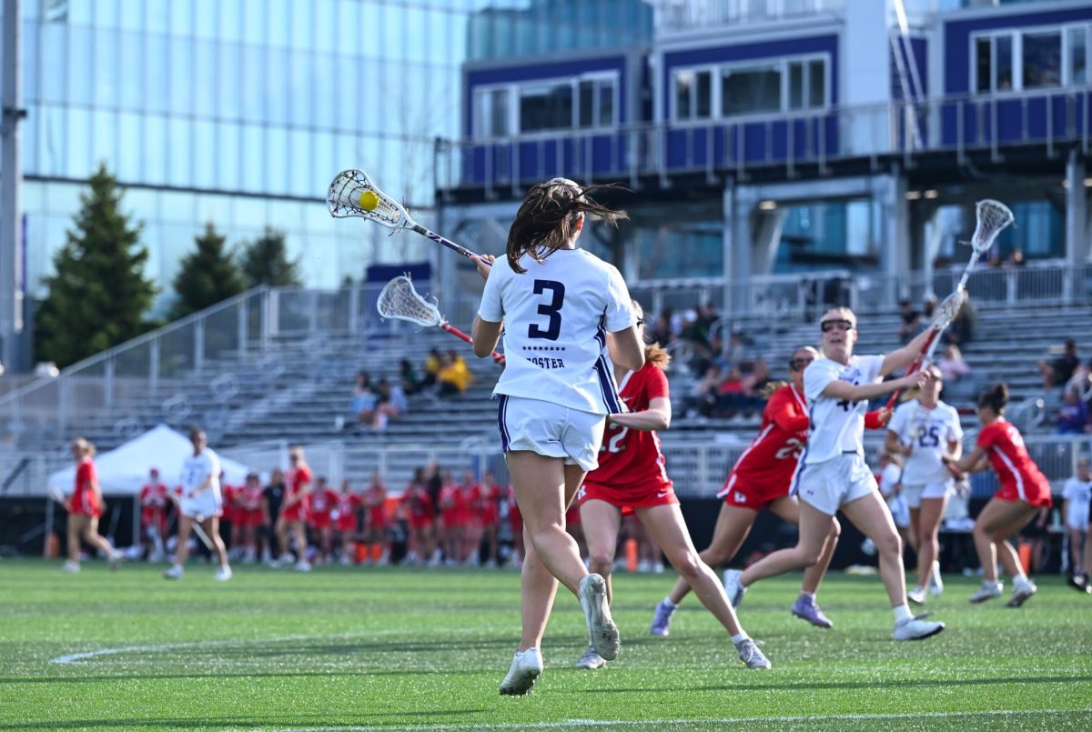 A woman in a white jersey with the ball in her stick faces away from the camera.