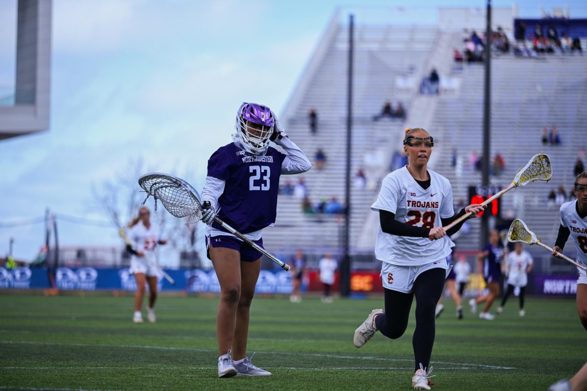 A player in a purple jersey and a player in a white jersey watch other lacrosse players.