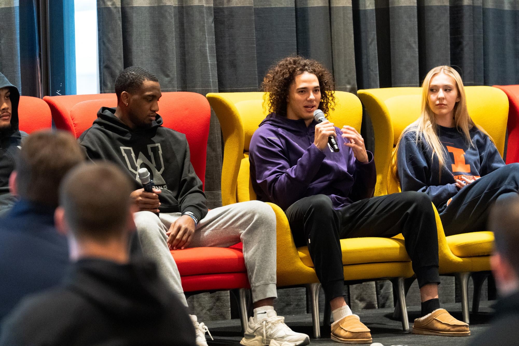Four student-athletes sit in colorful cushioned chairs on a stage, holding microphones and speaking to a crowd off-screen.