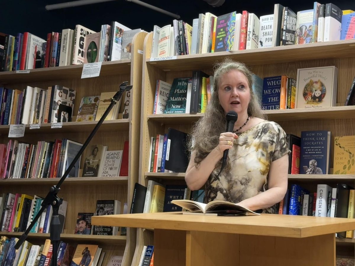 A person stands in front of a bookcase holding a microphone and book.