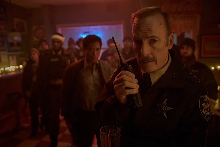 Bob Odenkirk talks into a walkie-talkie as a crowd of people stands behind him inside a dimly lit restaurant.